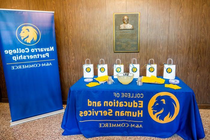 Goody bags and refreshments sit atop a table adorned with a blue and gold tablecloth next to an A&M-Commerce Navarro College Partnership flag banner.